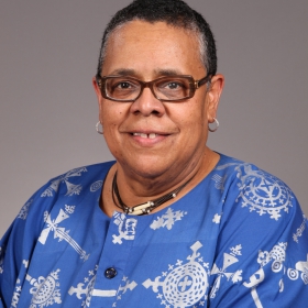 Margaret Woods, a woman with short hair and glasses, poses in front of a gray background. She is wearing a blue and white shirt.